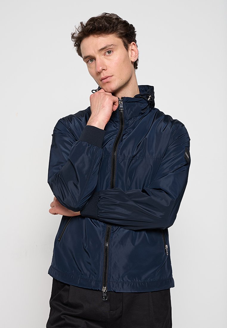 Navyblaue leichte Jacke mit hohem Kragen, Reißverschluss, elastischen Bündchen und Seitentaschen. Glatte, glänzende Textur und minimale Details.