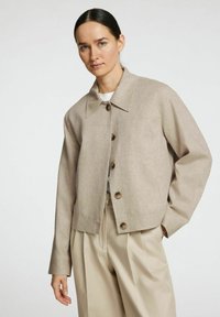 Chaqueta beige corta con cuello puntiagudo, grandes botones marrones y tejido texturizado; combinada con pantalones anchos beige.