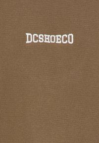 Ruskea puuvillakangas, jossa valkoisella kirjailtu teksti "DCSHOECO" keskellä. Yksinkertainen, teksturoitu pinta poimukuvioinnilla.