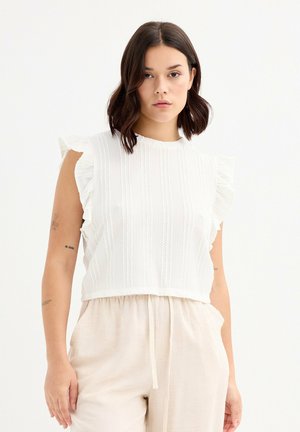 Top blanco cortado con mangas acampanadas, tejido texturizado con rayas verticales. Combinado con pantalones holgados de color claro.