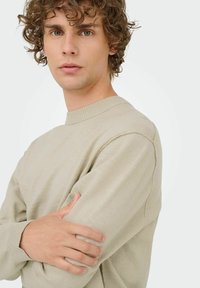 Beige gebreide trui met een ronde halslijn, voorzien van ribbelmanchetten en een subtiele textuur, gedragen door een persoon met krullend haar.