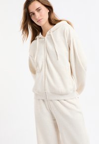 Sweat à capuche beige à fermeture éclair en tissu doux, doté d'une capuche, de cordons de serrage réglables et de poches latérales, accompagné d'un pantalon assorti.