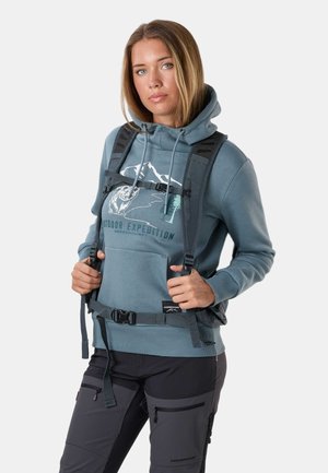 Junge Frau, die einen blauen Hoodie und Wanderhosen trägt, stellt die Rucksackriemen ein, bereit für eine Outdoor-Expedition.