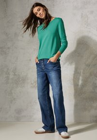 Grüner Pullover mit langen Ärmeln kombiniert mit blauen weiten Jeans, die einen Knopfverschluss und Fronttaschen haben. Weiße Sneakers runden das Outfit ab.