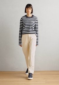 Stripet navy og hvit langermet topp, kremfargede bukser og mørke joggesko. Jevn tekstur og avslappet passform. Tremurgulv i bakgrunnen.