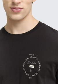 Čierne tričko s okrúhlym grafickým motívom obsahujúcim "Založené MMVII" a "Denim Design Studio Urban Crew" v bielej farbe písma a akcentoch.