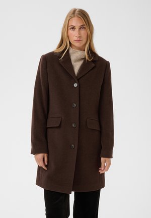 Femme blonde portant un manteau marron foncé boutonné par-dessus un pull beige à col montant sur un fond clair uni.