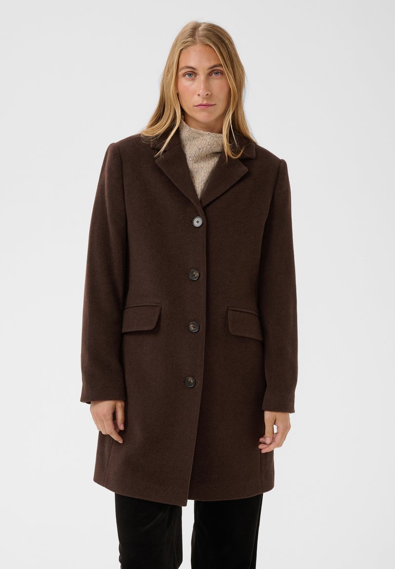Femme blonde portant un manteau marron foncé boutonné par-dessus un pull beige à col montant sur un fond clair uni.