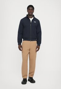 Navy bomber jacket with a zip closure, elastic cuffs, and light brown cargo pants with side pockets; worn with black lace-up shoes.