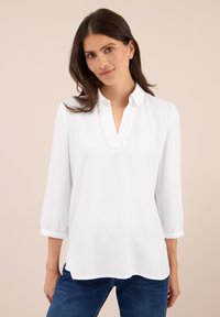 Femme aux cheveux bruns portant un blouse blanche à col en V avec des manches trois-quarts et un jean bleu, se tenant devant un fond beige.