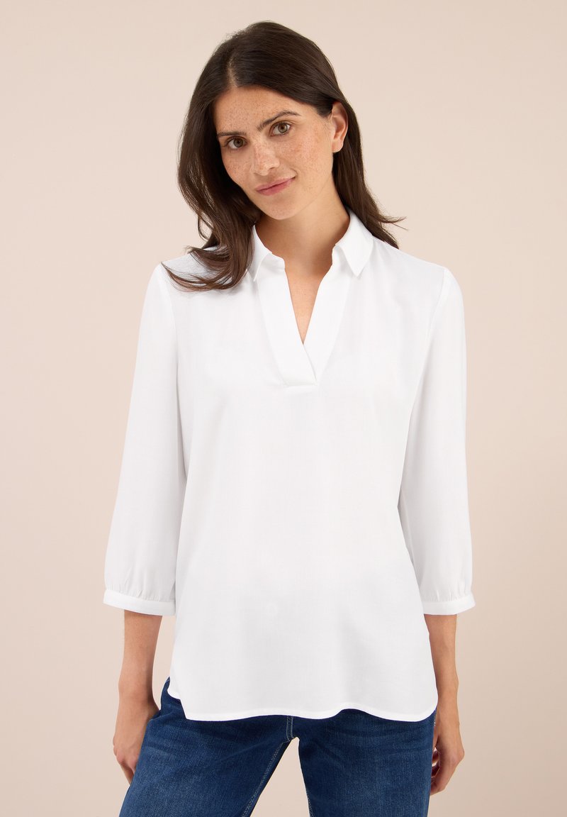 Femme aux cheveux bruns portant un blouse blanche à col en V avec des manches trois-quarts et un jean bleu, se tenant devant un fond beige.