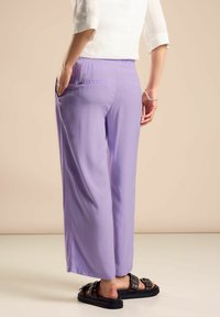 Lawendowe spodnie szerokie z miękkiej, gładkiej tkaniny, z elastycznym paskiem w talii i kieszeniami z tyłu, zestawione z czarnymi sandałami z akcentami metalowymi.