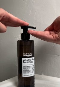 Bruine transparante fles met een zwarte pompdekking, gemerkt "Absolut Repair Molecular," gebruikt voor het doseren van een serumproduct. Handen zijn gepositioneerd om te pompen.