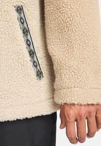 Beige fleecejakke med tekstureret overflade, med en sidelomme prydet med multifarvet geometrisk broderi og design med manchetteærmer.