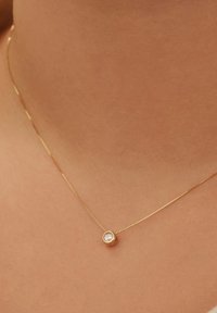 Goudkleurige ketting met een dunne schakel, voorzien van een ronde, heldere steen in een ronde gouden hanger, rustend op blote huid.