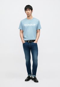 Απαλό γαλάζιο βαμβακερό t-shirt με το λογότυπο «DONDUP» σε λευκό, συνδυασμένο με σκούρο μπλε τζιν στενής γραμμής. Μια μαύρη ζώνη και μαύρα παπούτσια ολοκληρώνουν το σύνολο.