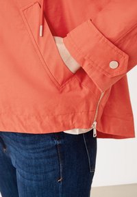 Orangene Regenjacke mit einem seitlichen Reißverschlussfach, Druckknöpfen an den Ärmeln und einem glatten, leicht strukturierten Stoff. Über dunklen Jeans getragen.