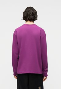 Άτομο με κοντά σκούρα μαλλιά, που φοράει μωβ πουκάμισο με μακριά μανίκια και μαύρο παντελόνι, στέκεται με την πλάτη προς το μέρος μας σε απλό φόντο.