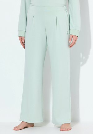Personne portant un pantalon large vert clair, debout pieds nus sur un fond blanc, les mains détendues le long du corps.