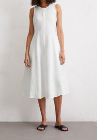 Sleeveless white linen dress with a flared shape, round neckline, and subtle texture. Paired with black slide sandals.