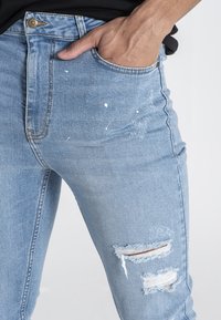 Ljusblå denimjeans med slim fit, med uppriven söm vid knäna och vita färgstänk. Standard femficksdesign med metallknapp.