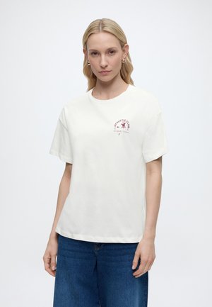 Femme blonde avec des boucles d'oreilles dorées portant un T-shirt blanc "Athletic Club" et un jean bleu, debout devant un fond uni.