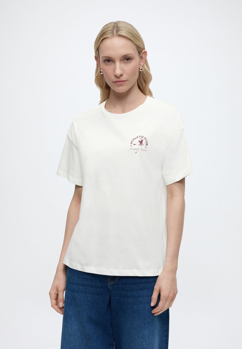 Ξανθιά γυναίκα με χρυσά σκουλαρίκια φορώντας λευκό μπλουζάκι "Athletic Club" και μπλε τζιν, στέκεται μπροστά σε απλό φόντο.