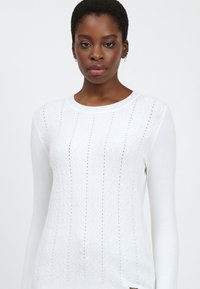 Suéter de punto blanco con un patrón acanalado y agujeros sutiles. Mangas largas y cuello redondo. Textura suave, diseño ajustado.
