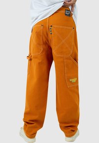 Pantalones cargo naranjas de algodón, con grandes bolsillos, costuras detalladas y un acento multicolor en la cinturilla. Incluye parche con el logo.
