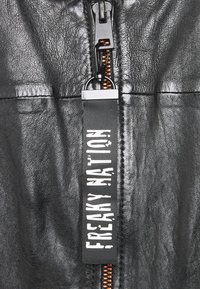 Černá kožená bunda s předním zipem a oranžovým podšívkou. Obsahuje látkovou etiketu s nápisem "FREAKY NATION" tučnými bílými písmeny.