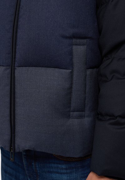 Navy blue puffer jacket with a textured fabric, contrasting lighter gray panel, and a zip closure. Features a side pocket detail.