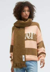 Woman with curly hair wearing a brown fringed scarf over a pink and brown striped sweater and light blue jeans, standing against a white background.