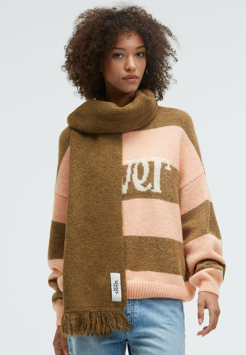 Woman with curly hair wearing a brown fringed scarf over a pink and brown striped sweater and light blue jeans, standing against a white background.