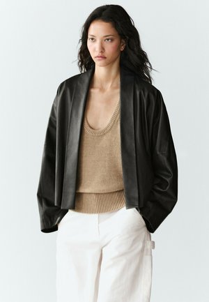 Femme aux longs cheveux foncés porte une veste en cuir noire sur un débardeur en maille beige et un pantalon blanc sur un fond clair uni.