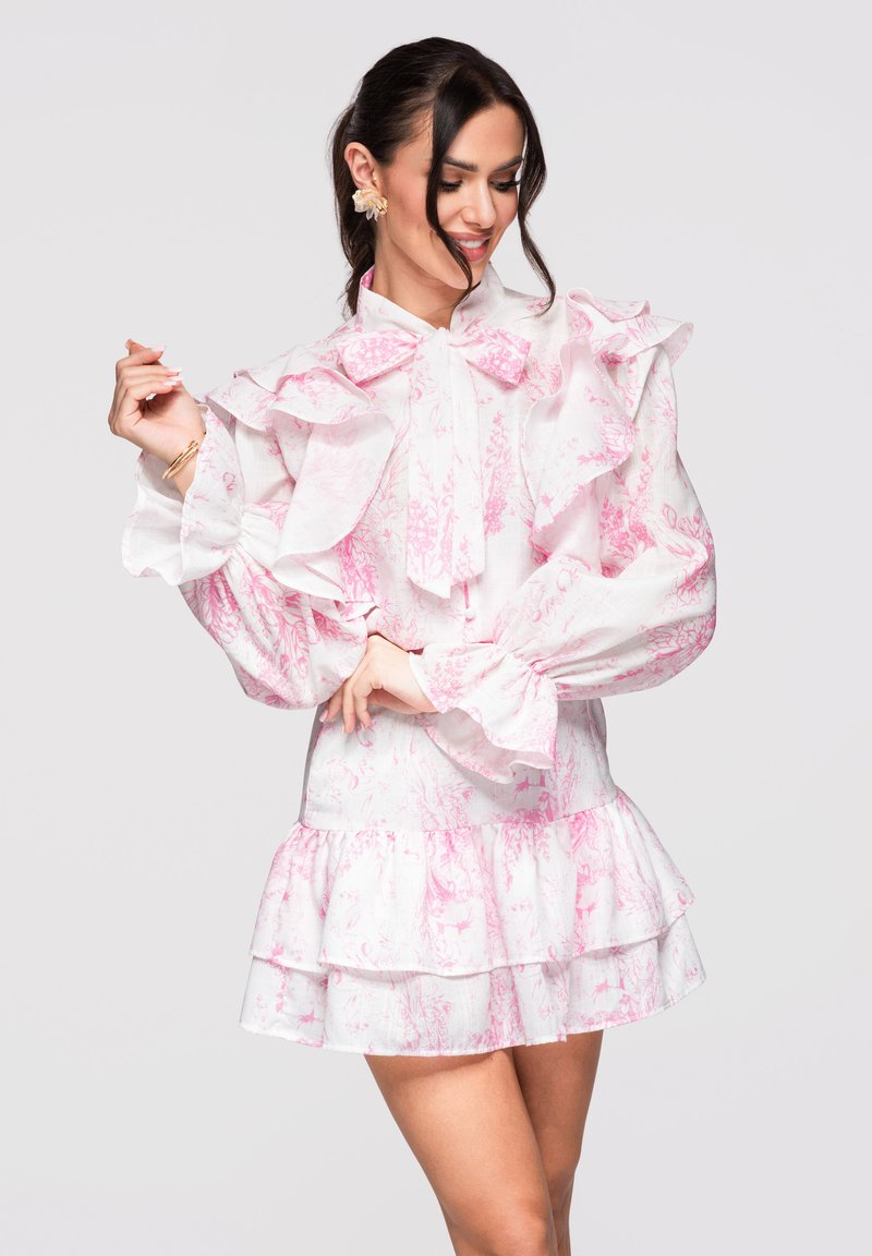 Femme portant une robe blanche à imprimé floral rose, manches volantées et jupe à volants, souriant et regardant vers le bas avec les mains en position.