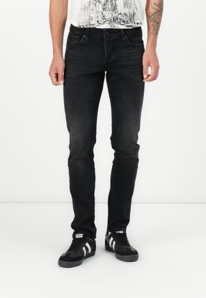 Mann trägt schwarze Slim-Fit-Jeans und schwarze Sneakers mit weißen Streifen, steht vor einem schlichten weißen Hintergrund.