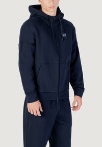 Marineblaue Zip-Hoodie aus weichem Stoff, mit Kapuze mit Kordelzug, Fronttaschen und Logo-Patch auf der Brust. Glatte Textur, moderner Schnitt.