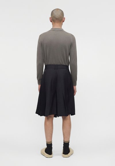Άτομο με ξυρισμένο κεφάλι που φοράει γκρι μπλούζα με μακριά μανίκια, μαύρη πλισέ φούστα, μαύρες κάλτσες και μπεζ παντόφλες, στέκεται γυρισμένο με την πλάτη.