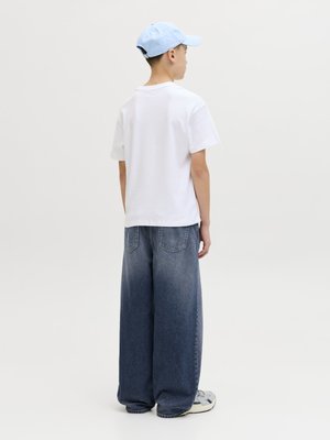 Nastolatek w białej koszulce, szerokich wyblakłych niebieskich dżinsach, jasnoniebieskiej czapce baseballówce i tenisówkach, stojący tyłem na jednolitym tle.