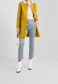 Geel wollen jas met een ronde kraag, witte blouse, gemusterde blauwe broek en witte enkellaarsjes, terwijl ze een kleine witte handtas vasthoudt.