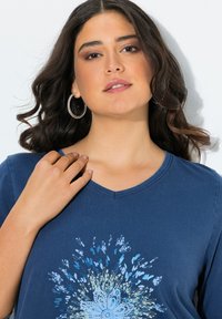 Junge Frau mit dunklem, welligem Haar, die große Creolenohrringe und ein blaues Oberteil mit floralem Burst-Design trägt und sanft ihre Schulter berührt.