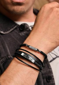 Bracciali intrecciati in pelle nera con accenti argentati e la scritta "SCORPIONS" incisa, indossati su un polso con texture della pelle visibile.