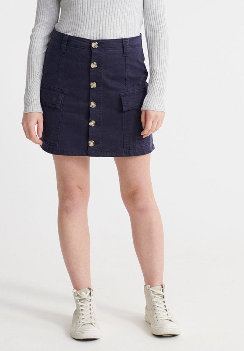 Marineblå denimnederdel med frontknaplukning, to sidelommer og en let buet kant. Stylet med en grå ribbet langærmet top.