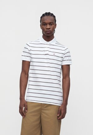 Junger Mann mit geflochtenem Haar, der ein weißes Poloshirt mit dünnen schwarzen horizontalen Streifen und beigen Hosen vor einem schlichten Hintergrund trägt.
