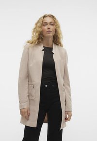 Blazer largo de color rosa claro con un acabado texturizado, que presenta dos bolsillos delanteros con cremalleras, diseño ajustado y cuello chal.