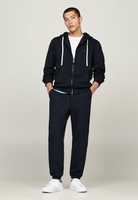 Haut de survêtement zippé en coton marine avec des cordons blancs, associé à un pantalon de survêtement assorti et des baskets blanches, présenté sur un fond neutre.