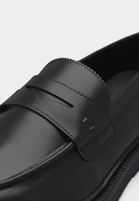 Μαύρο δερμάτινο μοκασίνι με ραμμένη λωρίδα για νόμισμα και λεπτομέρεια με κυματοειδές άκρο στο πάνω μέρος, εμφανιζόμενο σε κοντινή λήψη με λευκό φόντο.