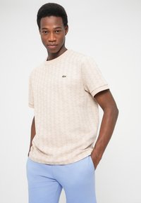 Giovane uomo che indossa una camicia beige a maniche corte con motivi e un piccolo logo verde a forma di coccodrillo, e pantaloni azzurri, con le mani nelle tasche.