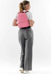 Zaino rosa realizzato in materiale testurizzato, con forma curva, manici superiori e una tasca con zip frontale, indossato con pantaloni larghi grigi.