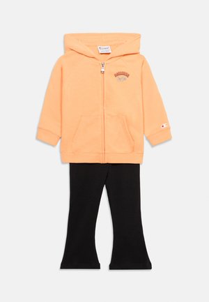 Jahodová mikina na zips s prednými vreckami a malým logom, kombinovaná s čiernymi rozšírenými nohavicami na bielom pozadí.